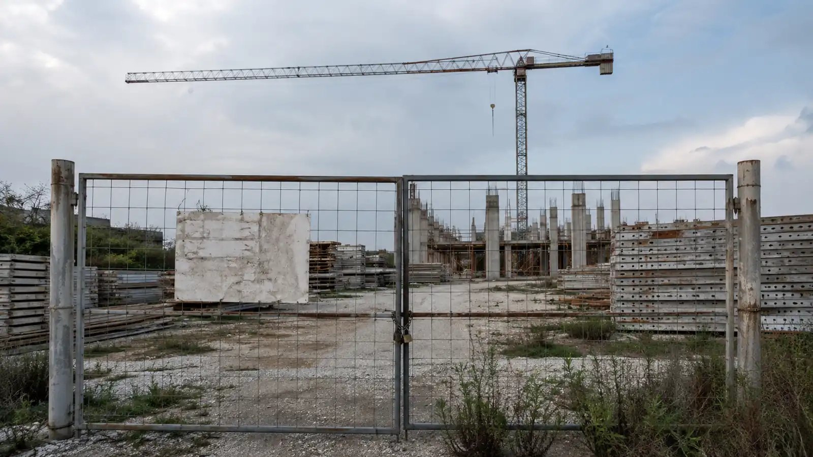 Cantiere edile fermo con gru e materiali accatastati, cancello chiuso con lucchetto e cartello di cantiere sbiadito