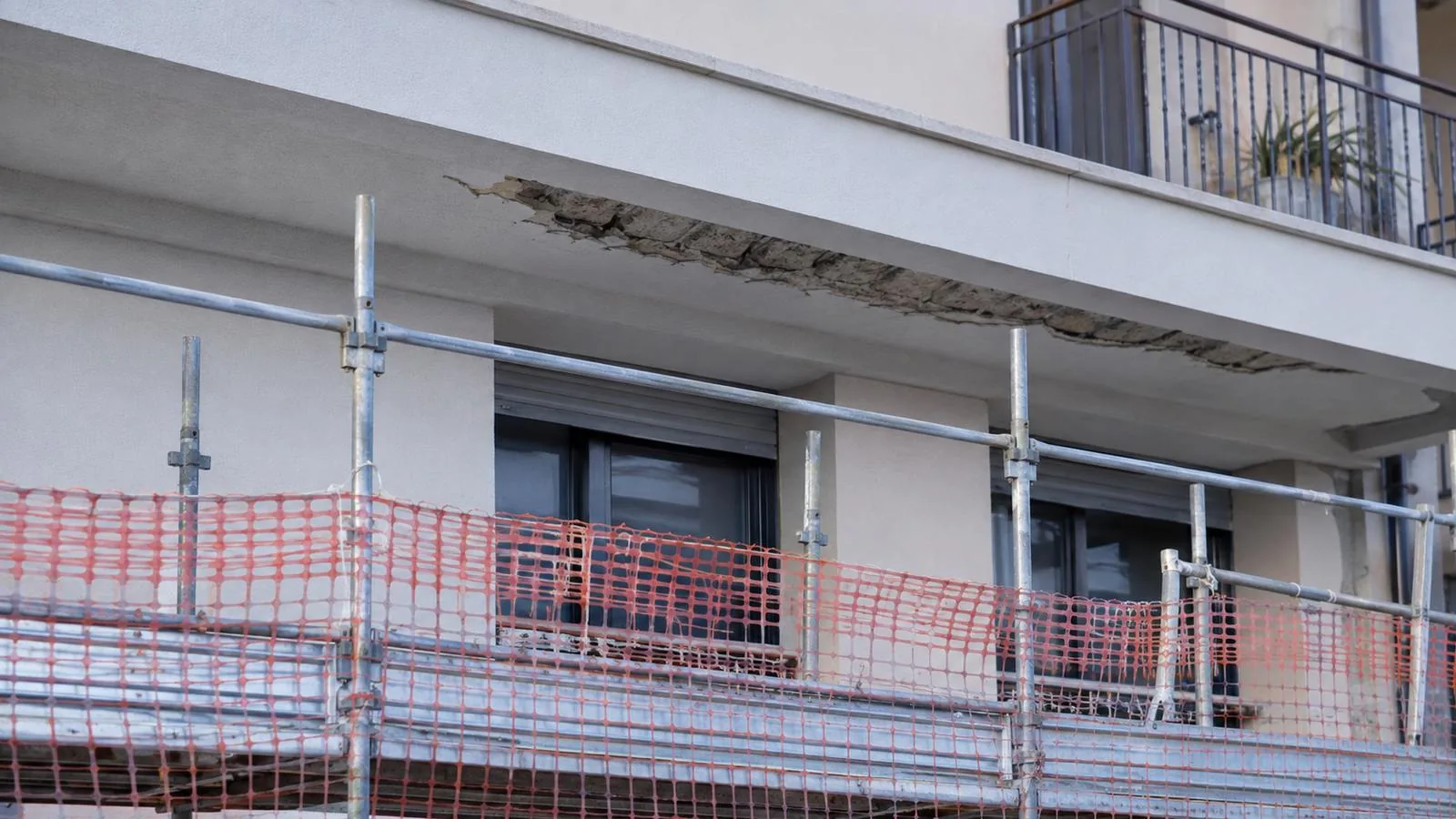 Facciata di una palazzina recente con transenne e rete di protezione sotto un cornicione con intonaco distaccato